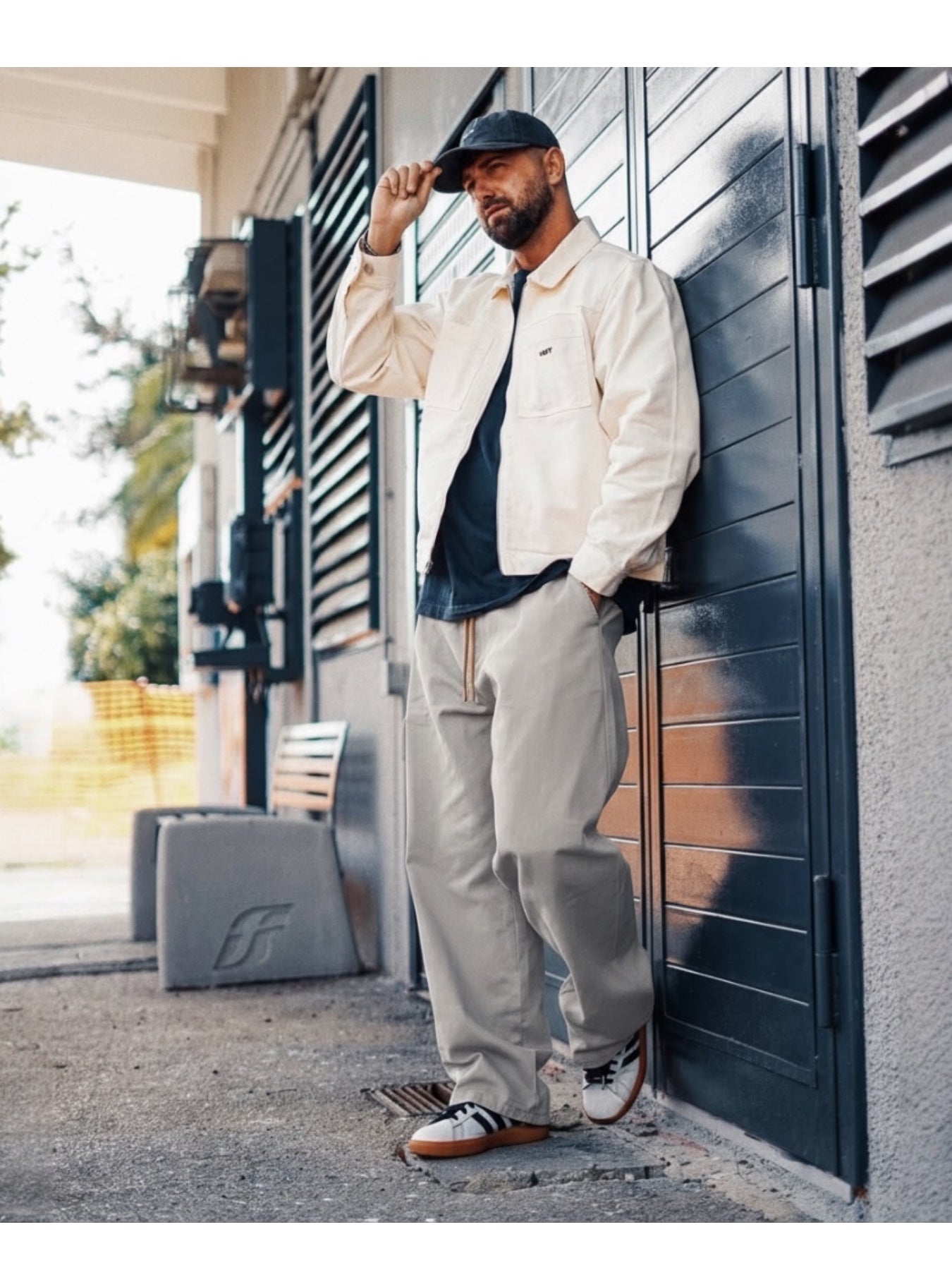 Manuel Store off-white cotton twill workwear jacket and drawstring pants worn by a man with a cap.